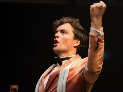 Opera student with shirt brown hair wear a pale pink suit and bow tie has one arm raised above their head whilst singing